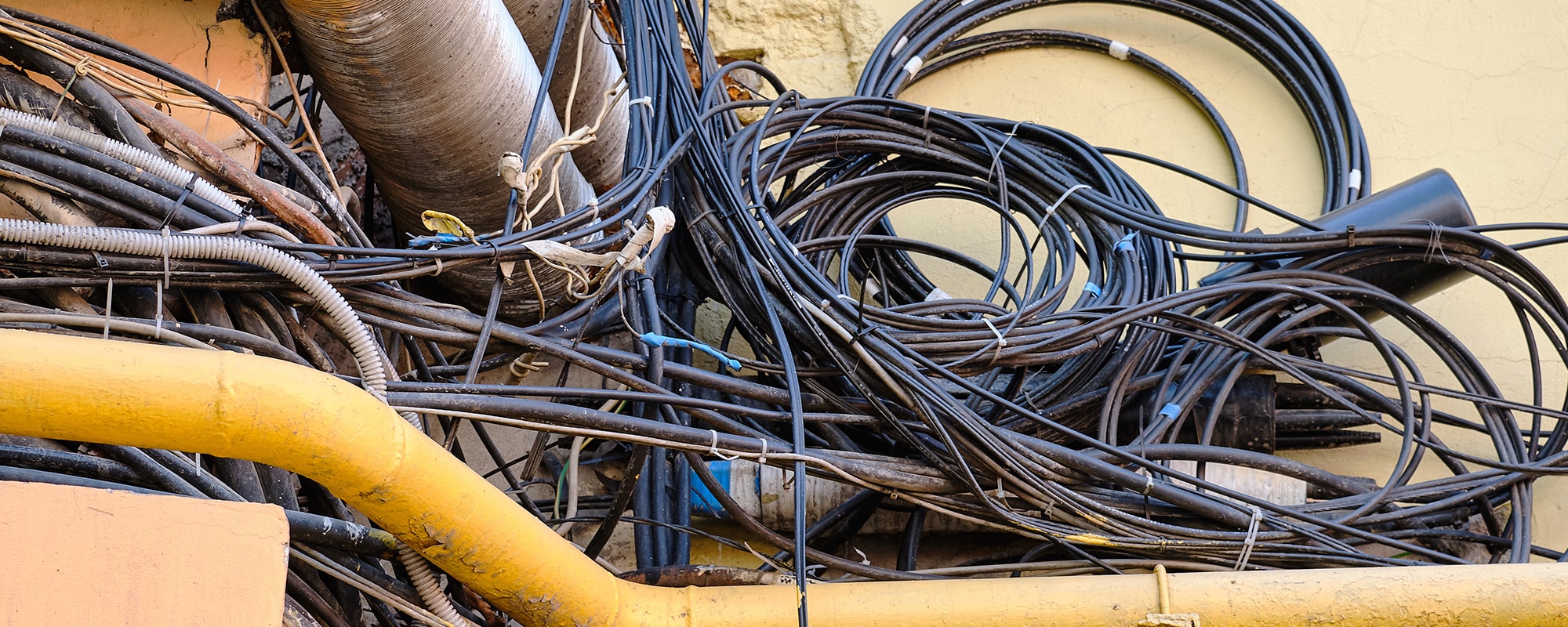 Bundles of black cables intertwined with yellow and silver colored, dusty, rusting pipes in an outdoor setting. 